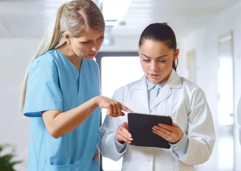 Two nurses, one of whom is a doctor, looking at a tablet in a hospital hallway.