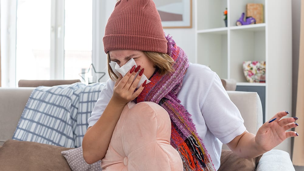 sick young woman sneezing in tissue during flu season on the couch in her home